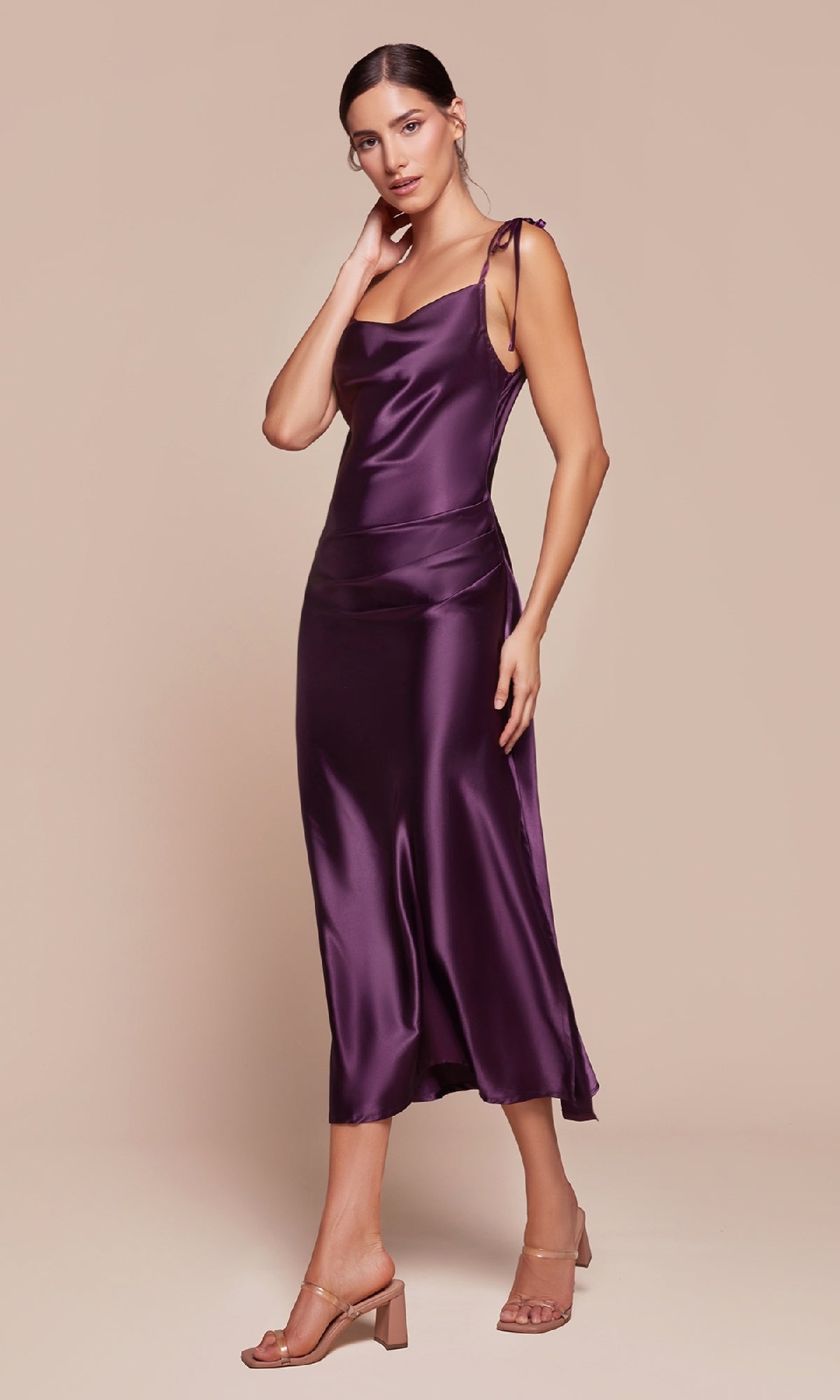 A woman models the Midi Wedding-Guest Slip Dress BD103, a purple semi-formal midi dress with tie straps, standing against a beige background with one hand on her neck and the other by her side.